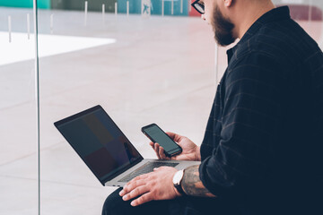 Cropped view of hipster bearded guy sharing media from laptop computer on digital smartphone using...