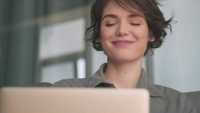 Smiling pensive young pretty woman using laptop computer and looking away while sitting on sofa at home