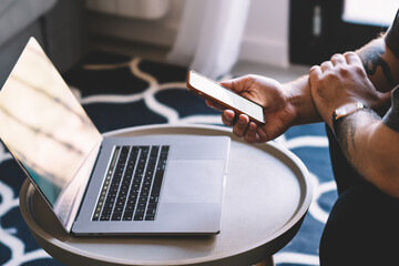 Cropped view of man's hand holding smartphone device near modern laptop computer and updating app.Hipster guy downloading media on digital cellular using high speed internet connection