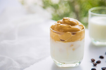 Close up a cup of Dalgona coffee with coffee beans next to a glass window on a white table with a cup of milk blurred and green nature background.