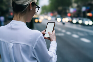 Cropped back view of hipster blogger holding smartphone device and touching with fingers on blank screen of device to search direction in app standing in urban setting on blurred background