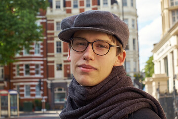Young casual handsome guy wearing brown glasses and a trendy gray ivy flat cap, glancing amused to the camera in an urban setting in London city.