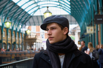 Young casual handsome guy wearing a brown scarf and a trendy gray ivy flat cap, visiting the interior of the former vegetable market of Covent Garden in London.