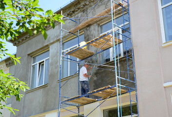 A building contractor on scaffoldings is plastering, rendering, coating, installing stucco on the facade of a residential building before painting the concrete exterior wall.