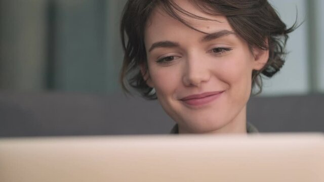 Happy pensive young pretty woman using laptop computer and looking away while sitting on sofa at home