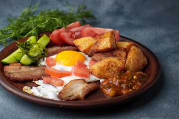 Breakfas on a clay platet, cucumbers, fried potatoes, fried egg, bacon, tomatoes and sauce, on blue concrete with herbs