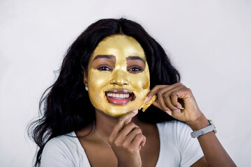 Skin care, beauty, spa. Pretty African woman with golden face mask. Close up of beautiful sexy girl taking off cosmetic peeling gold mask from healthy skin, posing to camera on white background