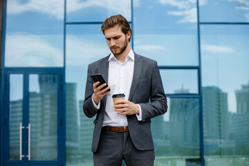 Fototapeta premium Well-dressed and successful young man in a stylish suit enjoys coffee and checks messages on his mobile phone outdoors in the background of a modern glass building.
