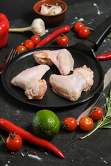 Chicken wings in a in a frying pan, fresh rosemary, fresh cherry tomatoes, fresh chili peppers, garlic, lime and sea salt on a black background.