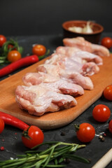 Chicken Leg in a wooden Cutting board, fresh rosemary, fresh cherry tomatoes, fresh chili peppers, garlic, lime and sea salt on a black background.