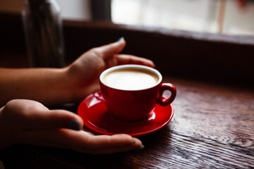 Cup of cappuccino in the hands of a young girl. Girl holding a red cup with coffee on a wooden background, hand plan and coffee. Top view. Place for text. Cappuccino with latte.