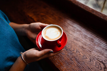 Cup of cappuccino in the hands of a young girl. Girl holding a red cup with coffee on a wooden background, hand plan and coffee. Top view. Place for text. Cappuccino with latte.