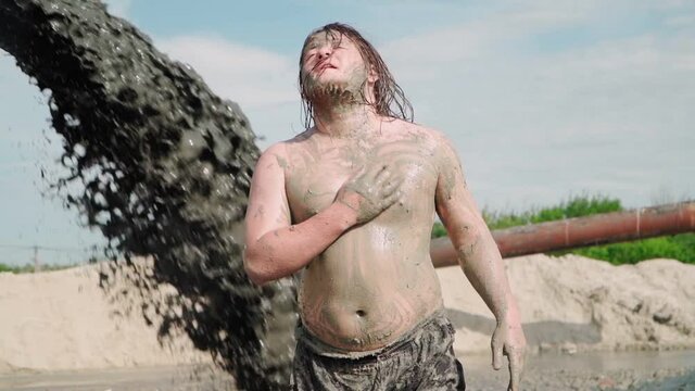 Funny Fat Man with Long Hair Bathes in the Mud. Against the background is pouring Black Dirty Water from the Pipe. Slow motion