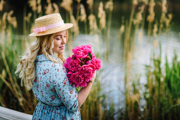 A young woman with a bouquet of pink peonies is resting on a pond beach. Girl in a blue dress and a hat with flowers. The concept of outdoor recreation. Summer holidays.