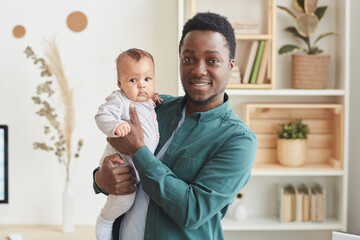 Fototapeta premium Waist up portrait of young African-American man smiling happily at camera while posing with cute baby in home interior, copy space