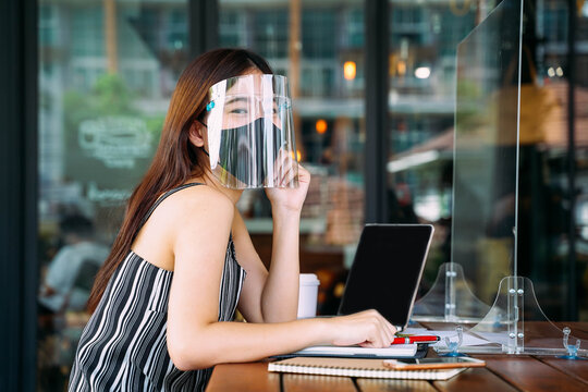 Young 20s Asian beautiful woman wearing a protective face shield and mask with partition in cafe restaurant. While using computer laptop and mobile phone outside. - Corona Virus prevention concept.