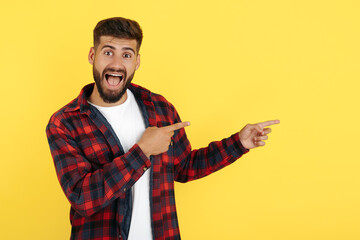 Delighted bearded hipster young man in plaid shirt point fingers away over yellow background. Look in camera.