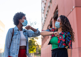 Young and friendly african girl and caucasian girl keeping social distance, greeting each other by bumping elbows instead of hugs, kisses or handshaking, wearing mask for prevent coronavirus infection © MandriaPix