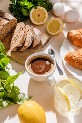 fresh croissants, bread, greenery, eggs, lemon water and cup of coffee for breakfast on grey table