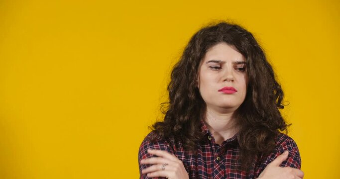 Close-up attractive plus-size model posing. A curly-haired brunette on bright yellow studio background demonstrating different emotions. Girl is offended, angry with her arms crossed and frowning.
