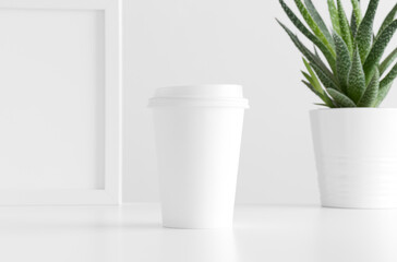 Coffee paper cup mockup with a cactus in a pot and a frame on a white table.