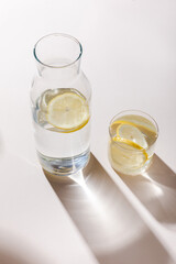 fresh water with lemon slices in carafe and glass on white table with shadows