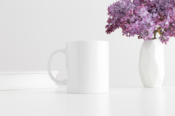 White mug mockup with a book and a bouquet of lilac in a vase on a white table.