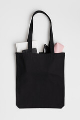 Black tote bag mockup with a notebook, book, glasses and case on a white table.