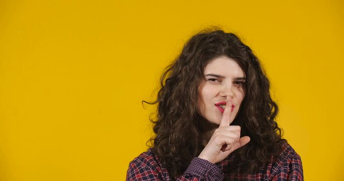 Close-up attractive plus-size model posing. Curly-haired brunette on bright yellow studio background demonstrating different emotions. Girl bringing index finger to her lips and asking don&rsquo;t speak