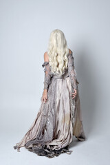 full length portrait of blonde girl wearing long torn old wedding dress.  standing pose with back to the camera on a studio background.