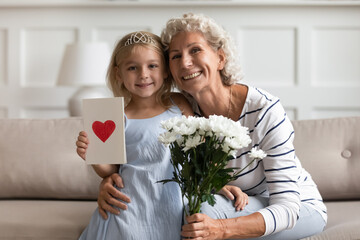 Little granddaughter with postcard red painted heart on it old grandmother holding white flowers...