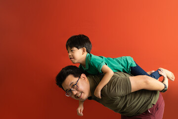 Asian father giving son ride on the back with a red background. Portrait of a happy father giving son piggyback a ride on his shoulders and looking up.