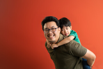 Asian father giving son ride on the back with a red background. Portrait of a happy father giving son piggyback a ride on his shoulders and looking up.