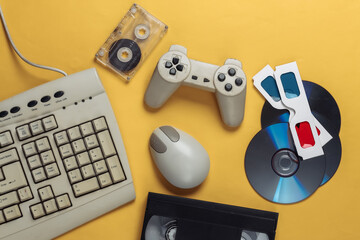Retro entertainment. Old-fashioned keyboard, pc mouse, compact discs, gamepad, anaglyph glasses, audio and video cassette on yellow background. Top view. Flat lay