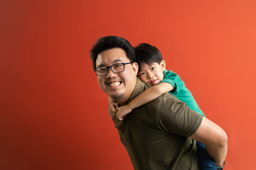 Asian father giving son ride on the back with a red background. Portrait of a happy father giving son piggyback a ride on his shoulders and looking up.