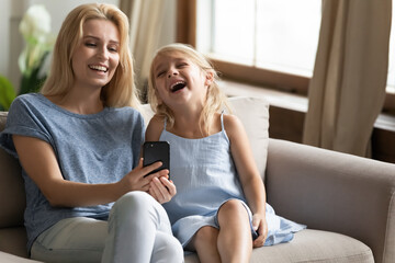 Obraz premium Happy daughter and cheery mom sit on couch use internet entertainment amusements on-line, watching cool funny prank videos on smartphone and laughing, modern tech and juniors, parental control concept