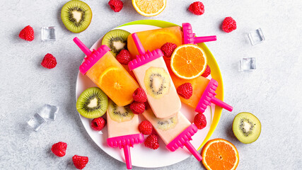 Fototapeta na wymiar Summer refreshing homemade popsicles with oranges and kiwi. Healthy snack with fruits and frozen juice for summer heat on concrete table top, flat lay, copy space