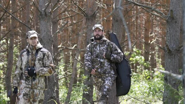 Two hunter men in camouflage clothes with guns walking through forest during hunting season. Man hunter outdoor in forest hunting. Male Tourists. Slow motion. Summer hiking in wilderness area.