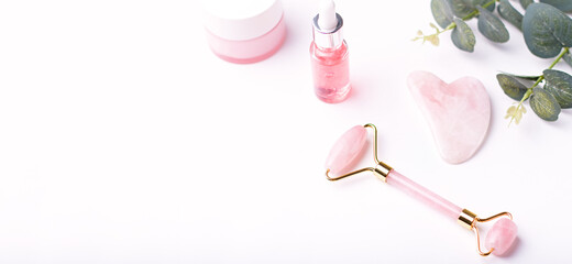 Pink hyaluronic acid serum, rose quartz roller, gua sha and moisturizing face cream on white table top. Anti aging cosmetics and tools on white, copy space, top view, web banner