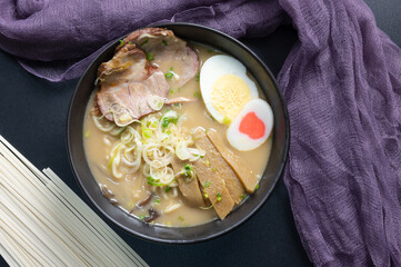 Delicious tonkotsu ramen, Japanese noodle in pork Bone based soup topped with chashu pork, boiled egg, pickled bamboo shoot and spring onions. top view on black background Decorated with purple napkin