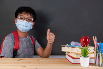 School boy wearing face mask thumbs up over blackboard with copy space. Safe back to school during...