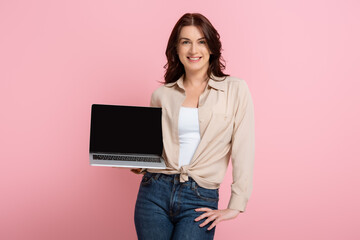 Attractive smiling woman with hand on hip showing laptop isolated on pink, concept of body positive