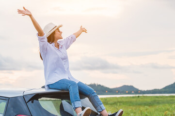 Happy Asian woman spread arms on car roof under sunset at seaside with nature background during travel in holiday