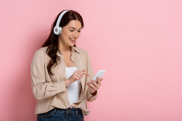 Positive woman in headphones pointing with finger at smartphone on pink background