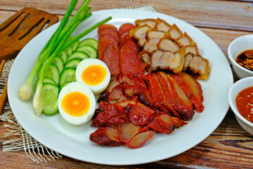 Barbecued red pork, crispy pork belly, Chinese sausage and boiled egg served with spring onion and cucumber slices having red sauce and pickled chilli aside on wooden table.