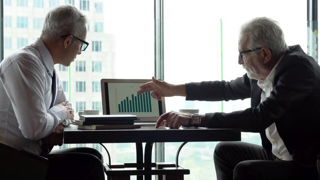 two senior businessman in meeting pointing graph in laptop analyzing  data information . old man ceo discussing with client in cafe. serious mature manager talking by window over cityscape luxury