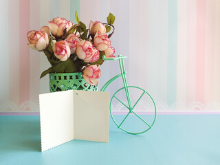 Against the background of striped wallpaper in pink pastel colors, there is a decorative bicycle with flowers. In the foreground is an open card for congratulations. Space for text. Exquisite backgrou