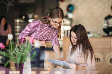 Hospitable waitress help to female customer what to choose something from menu. Smiled woman sitting in cafe