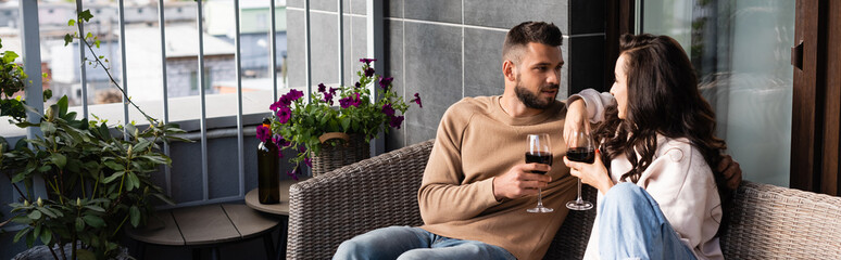 panoramic concept of handsome man and attractive woman sitting on outdoor sofa and holding wine glasses while looking at each other