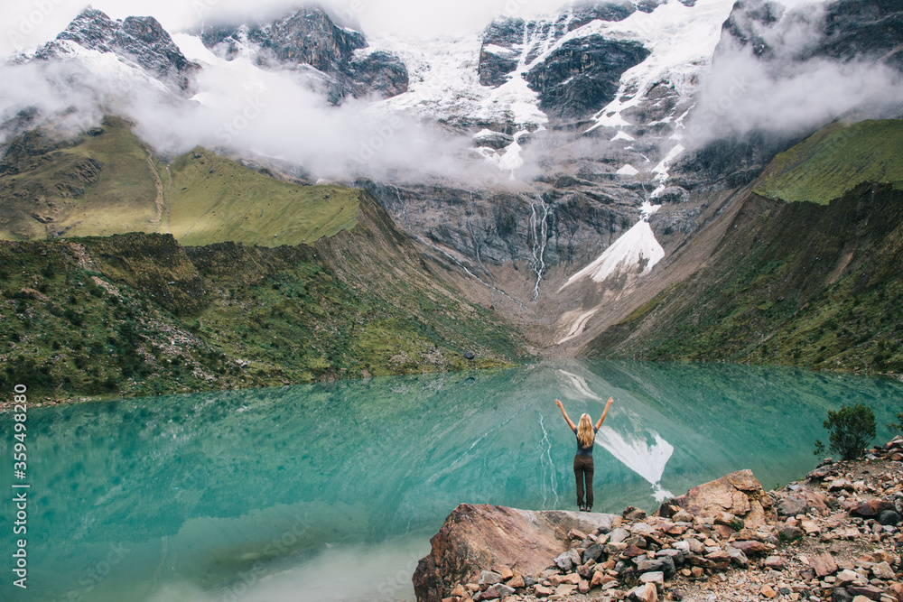 Wall mural Back view of female tourist holding hands up enjoying freedom and amazing scenery of mountains covered snows and clouds.Young woman hiker standing on rock of hill admiring lake and nature during trip